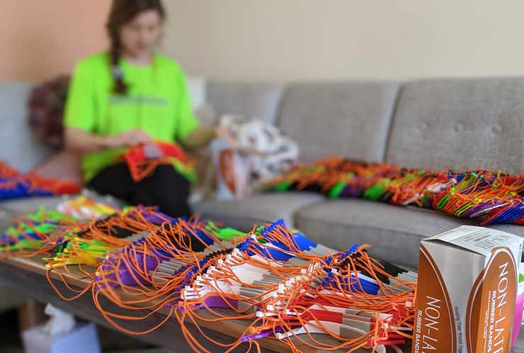 A woman is sitting among hundreds of personal protective face shields as she continues to assembled straps to the forehead frames.